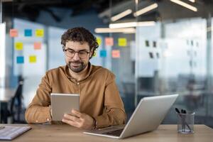 konzentriert männlich Geschäft Fachmann mit ein Tablette und Laptop beim ein modern Arbeitsplatz mit klebrig Anmerkungen im das Hintergrund. foto