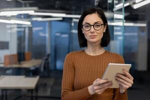 ein Fachmann Frau selbstbewusst hält ein Tablette im ein modern Büro Umfeld, Anzeigen Haltung und Sachverstand. foto