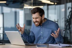 ein wütend Geschäftsmann ist Gefühl frustriert mit Technologie, zeigen Stress und Aggression gegenüber seine Laptop im ein Büro Umfeld. foto