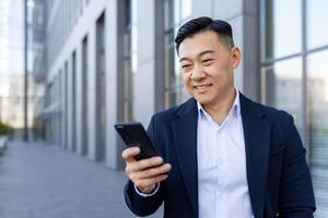 asiatisch lächelnd jung Mann Gehen Nieder das Straße in der Nähe von ein Büro Gebäude und mit ein Handy, Mobiltelefon Telefon. foto