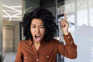 afrikanisch amerikanisch Frau zeigen nach oben und schreien, porträtieren Frustration oder ein dringend Erläuterung während ein Anruf im ein modern Büro. foto