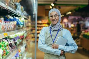 ein jung Frau, ein Supermarkt Arbeiter im ein Kopftuch. er steht in der Nähe von das Zähler mit Waren, lächelt foto