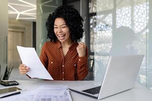 erfolgreich Financier Frau zufrieden mit Ergebnisse ,Überprüfung Bericht Geschäftsfrau beim Arbeitsplatz, glücklich halten Hand oben triumphierend Gewinner Geste afrikanisch amerikanisch Boss hinter Papier Arbeit mit Notizbuch foto