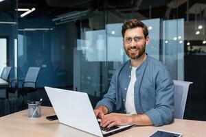 Porträt von reifen Geschäftsmann Innerhalb modern Büro, Senior Mann im Hemd lächelnd und suchen beim Kamera, Arbeiter mit Laptop beim arbeiten. foto