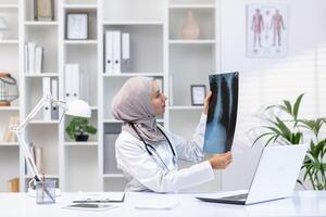 konzentriert Muslim Frau Arzt mit Stethoskop Überprüfung Röntgen von Lunge während Sitzung beim Krankenhaus Kabinett mit Laptop. qualifiziert Lungenarzt im Weiß medizinisch Uniform Herstellung Diagnose zum geduldig. foto