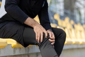 Nahansicht von ein männlich Athlet im Sportbekleidung Sitzung auf Stadion steht, umklammern seine Knie im leichte Schmerzen, vorbildlich Sport Verletzung. foto