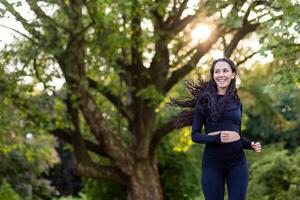 jung motiviert und froh Frau Joggen im das Morgen im ein Öffentlichkeit Park unter Bäume, spanisch Frau mit lockig Haar, schlank im ein Trainingsanzug. foto