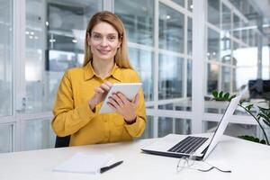 Porträt von ein erfolgreich lächelnd Frau Innerhalb das Büro beim das Arbeitsplatz, ein Financier Geschäftsfrau ist suchen beim das Kamera, mit ein Tablette Computer auf ein Laptop beim arbeiten. foto