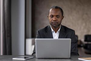 ernst konzentriert männlich Geschäftsmann Arbeiten Innerhalb Büro mit Laptop, reifen erfahren afrikanisch amerikanisch Mann Tippen auf Klaviatur, Denken lesen von Monitor. foto