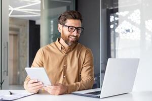 Porträt von ein jung Architekt, Designer Arbeiten im das Büro beim ein Laptop, reden auf ein Forderung, halten ein Tablette im seine Hände. foto