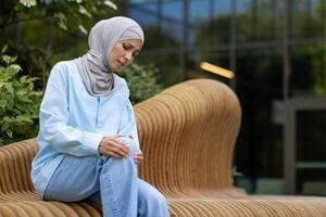 Muslim weiblich Sitzung draußen und erziehen Bein gebogen im Knie mit Leiden Ausdruck auf Hintergrund von modern Gebäude. krank Frau gegenüber Trauma Konsequenzen und Reiben verletzt Platz mit beide Hände. foto