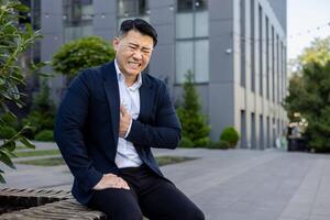 ein jung asiatisch Geschäftsmann sitzt auf ein Bank draußen ein Büro Gebäude. halten seine Truhe mit seine Hand und Leiden von Schmerz. foto