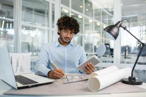 konzentriert jung männlich Architekt mit lockig Haar Arbeiten auf architektonisch Pläne im ein hell, modern Büro mit Digital Tablette und Laptop. foto