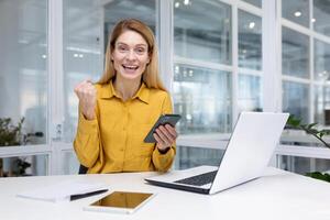 glücklich blond Geschäft Frau Sitzung beim ein Schreibtisch im ein hell Büro, halten ein Smartphone im ihr Hand, genießen gewinnen das Lotterie, lesen gut Nachricht, bekommen ein Förderung, suchen beim Kamera. foto