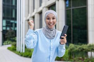 aufgeregt Muslim weiblich Jubel mit Handy, Mobiltelefon Telefon im angehoben Hände während Gehen Nieder Geschäft Bezirk. emotional Frau Empfang unerwartet angenehm Benachrichtigung Über Gehalt Transfer im Bankwesen App. foto