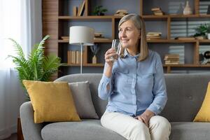 optimistisch Alten Frau mit grau Haar Sitzung auf Couch mit Kissen während bringen Glas von rein Wasser zu Mund. kaukasisch weiblich gekleidet im Licht Kleider Aufrechterhaltung gesund Gewohnheiten beim heim. foto