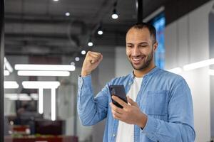 froh indisch Mann im ein beiläufig Denim Hemd ist feiern ein erfolgreich Geschäft Deal oder Leistung während mit ein Smartphone im ein Büro Einstellung. foto