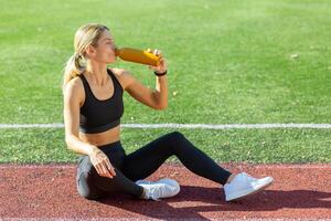 passen weiblich Athlet Sitzung auf ein Spur Feld nehmen ein brechen zu trinken von ein Flasche gefüllt mit ein Sport trinken auf ein sonnig Tag. foto