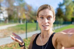 weiblich Athlet nach Joggen im Stadion reden mit freunde und Aufzeichnung online Blog, reifen blond mit Kopfhörer suchen beim Smartphone Kamera lächelnd, Anruf mit Kopfhörer. foto