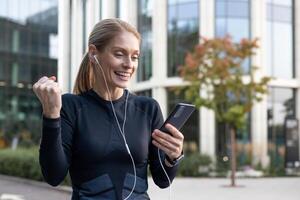 ein heiter Frau feiert ein Moment von Sieg oder gut Nachrichten draußen, Hören zu Musik- mit Kopfhörer von ihr Smartphone. das Hintergrund Vitrinen städtisch Grün und modern die Architektur, hervorrufen ein Sinn von ein Arbeitsleben Gleichgewicht. foto