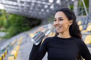 lächelnd sportlich Frau bleiben hydratisiert mit ein Wasser Flasche beim ein Sport Stadion. Gesundheit und Fitness Lebensstil Konzept gefangen draußen. foto