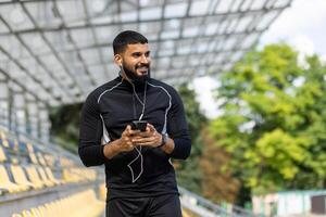ein lächelnd, bärtig Mann tragen Sportbekleidung und Kopfhörer hält ein Smartphone während Stehen beim ein draussen Stadion. foto