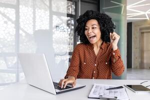 froh erfolgreich zufrieden afrikanisch amerikanisch Geschäftsfrau feiern großartig Leistung Ergebnisse beim Arbeitsplatz Innerhalb Büro, weiblich Arbeiter feiern halten Hand oben lächelnd mit Laptop. foto