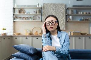 Depression. müde und nachdenklich jung asiatisch Frau Sitzung auf Sofa beim heim, halten Hand zu Kopf, suchen fokussiert. foto