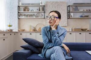 jung schön asiatisch Frau Denken im Brille Sitzung auf Sofa beim Zuhause im Leben Zimmer foto