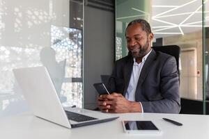 reifen erfahren und erfolgreich afrikanisch amerikanisch Boss beim Arbeitsplatz Innerhalb Büro, Senior zufrieden mit Ergebnisse Geschäftsmann mit Telefon, lesen Nachrichten von online App auf Smartphone. foto