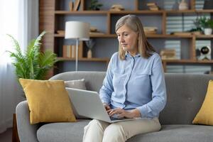 ein reifen Frau, konzentriert und beschäftigt, Verwendet ein Laptop während Sitzung auf ein Sofa im ein gut dekoriert Leben Zimmer, vorschlagen ein Mischung von Komfort und Produktivität. foto