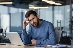 besorgt männlich Fachmann Erfahrungen Stress beim arbeiten, Sitzung beim seine Schreibtisch mit ein Laptop im ein zeitgenössisch Büro Einstellung. foto