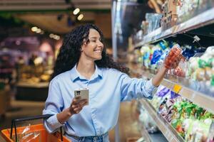 Frau Käufer glücklich mit Telefon im Lebensmittelgeschäft Abteilung Surfen Einkaufen aufführen im online Anwendung, wählen Gemüse Tomaten im Lebensmittelgeschäft Abteilung foto