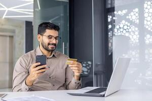 Fachmann männlich Unternehmer mit Smartphone und Anerkennung Karte zum online Transaktion beim seine modern Arbeitsplatz. foto