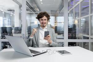 begeistert jung Erwachsene Arbeiten beim seine Büro Schreibtisch, zeigen Sieg Geste mit ein Smartphone im Hand, personifizierend Erfolg und Vertrauen im ein korporativ Umfeld. foto