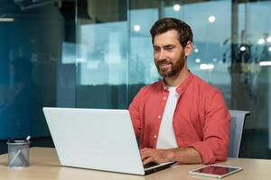 erfolgreich Geschäftsmann im rot Hemd glücklich Arbeiten mit Laptop Innerhalb Büro, reifen Mann mit Bart beim Arbeitsplatz Tippen auf Tastatur lächelnd zufrieden mit Arbeit Ergebnisse und Leistung. foto