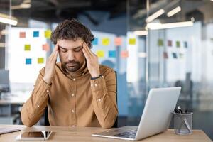 ein männlich Fachmann im ein braun Hemd fühlt sich Schmerz, halten seine Kopf im Not während Sitzung beim seine Büro Schreibtisch, Anzeige Stress oder Migräne. foto