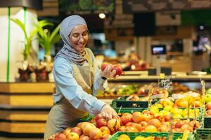 weiblich Verkäufer im Hijab Surfen und Überprüfung Äpfel im Supermarkt, Frau im Schürze lächelnd beim Arbeit im Geschäft im Obst und Gemüse Abteilung foto