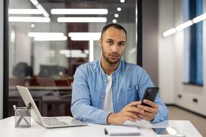 Porträt von ein ernst Latein amerikanisch Mann Sitzung im das Büro beim das Schreibtisch, halten ein Telefon im seine Hände und suchen beim das Kamera. foto