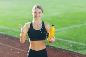 Porträt von glücklich Frau im Sportbekleidung, blond suchen beim Kamera und zeigen Daumen hoch, Sportlerin im Stadion tun Übung und Fitness, Hören zu online Radio und Musik, mit Flasche von Wasser. foto