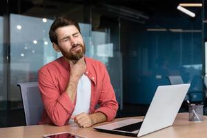 ein Mann mit ein wund Kehle funktioniert Innerhalb das Büro, ein Geschäftsmann beim Arbeit hat ein wund Kehle Grippe und kalt, Verwendet ein Laptop beim arbeiten. foto