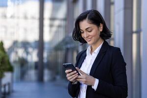 ein jung schön Geschäft Frau ist Gehen draußen das Büro Gebäude, ein Frau im Geschäft Kleider ist lächelnd, mit ein Anwendung auf das Telefon, Surfen das Internet, Tippen ein Botschaft. foto