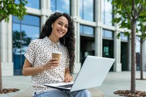 heiter spanisch Frau Sitzung draußen mit ein Laptop und Kaffee, genießen ein sonnig Tag beim Arbeit umgeben durch städtisch Grün. foto