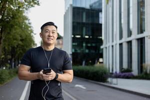 ein asiatisch jung lächelnd Mann ist tun ein Morgen Joggen auf das Stadt Straßen. Stehen im Sportbekleidung und Kopfhörer, halten ein Telefon. foto