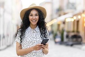 Porträt von ein jung schön Latein amerikanisch Frau Gehen im das Abend Stadt, halten ein Telefon, lächelnd und suchen beim das Kamera. foto