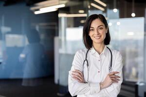 Fachmann weiblich medizinisch Praktiker lächelnd selbstbewusst im ein Klinik Einstellung, Waffen gekreuzt, tragen ein Labor Mantel und Stethoskop. foto