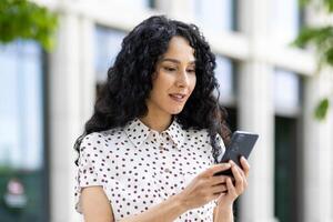 ein jung schön Frau Spaziergänge durch das Stadt mit ein Telefon im ihr Hände, lächelt zufrieden, Verwendet ein Smartphone Anwendung, durchsucht Internet Seiten, Typen ein Text Nachricht, Sozial Netzwerke. foto