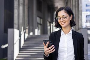 elegant Geschäftsfrau im ein passen lächelnd während suchen beim ihr Telefon, Stehen auf Schritte von ein modern Büro Gebäude. foto