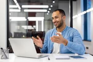 ein lächelnd jung Muslim Mann Sitzung im das Büro beim ein Schreibtisch im Vorderseite von ein Laptop und reden auf ein Forderung. foto