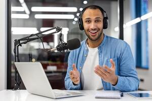 ein indisch Mann im beiläufig Kleidung ist Podcasting im ein modern Leben Zimmer, Erstellen Inhalt mit ein Fachmann Mikrofon und Laptop. foto